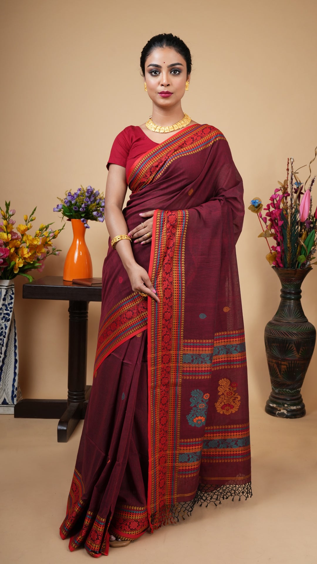 Short maroon Tangail soft cotton saree with floral motif border in red, orange, beige, and sea green. Butis and pallu carry the same motif for a balanced traditional look.