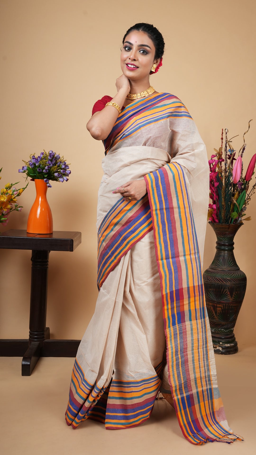Full-length photo of a woman in a light beige cotton Tangail saree with a multi-colored striped border, styled with gold jewelry.