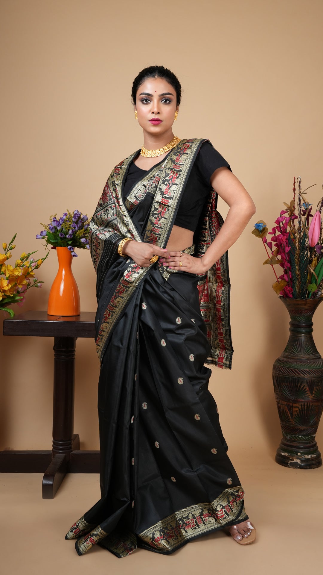 A woman posing in a black silk Baluchari saree with a rich red and beige woven border and small butis across the body.