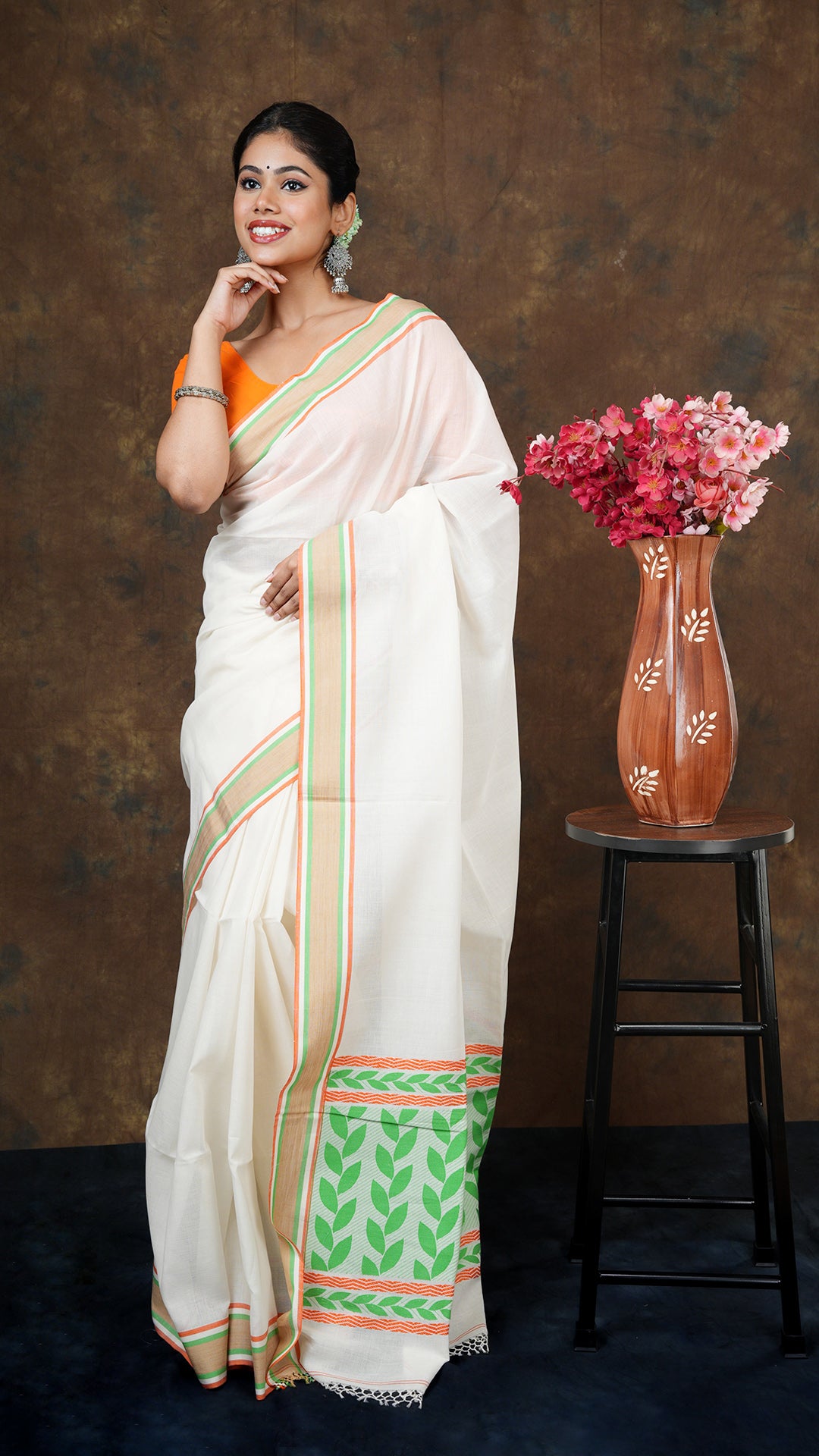 Off white tangail saree with beize, green, and orange border. Floral pallu in green and orange. Includes 0.8m running blouse. 5.5 meters.