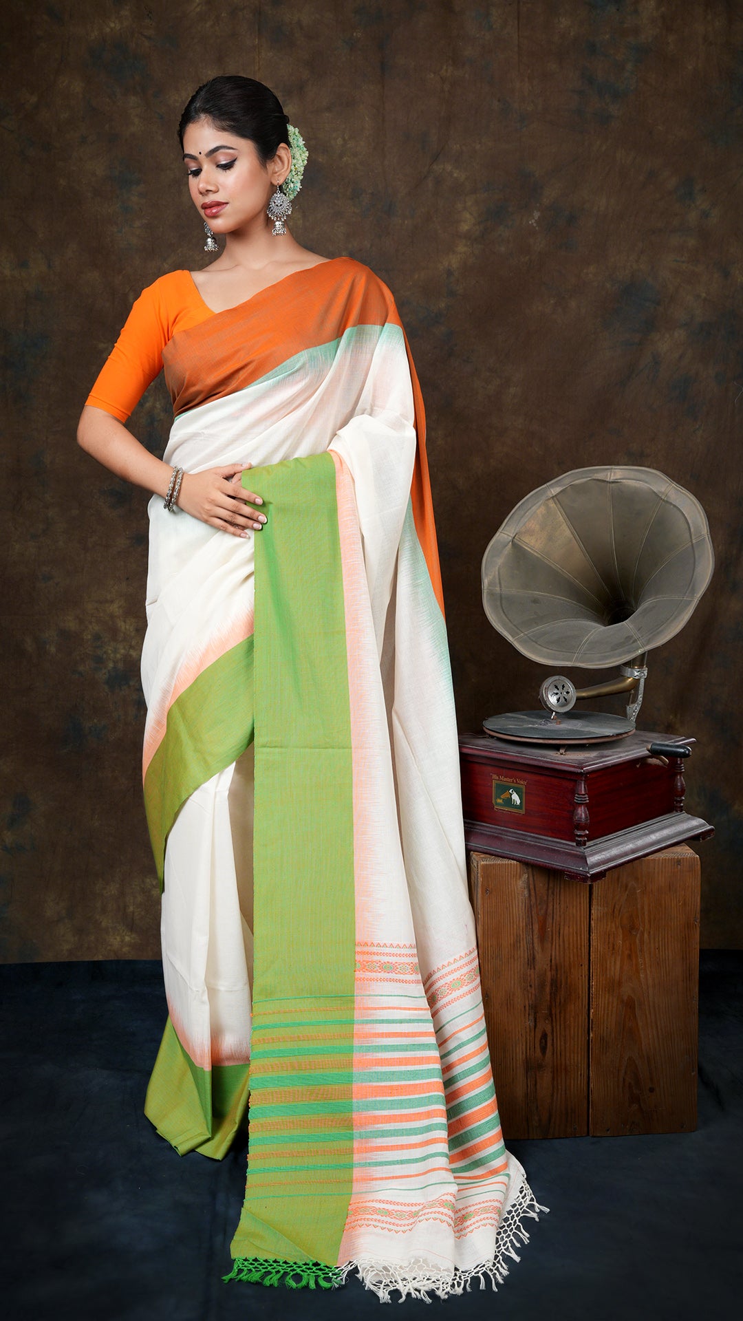 Off white tangail saree with flat orange and green borders. Pallu in orange-green weave. Includes 0.8m running blouse. 5.5 meters.