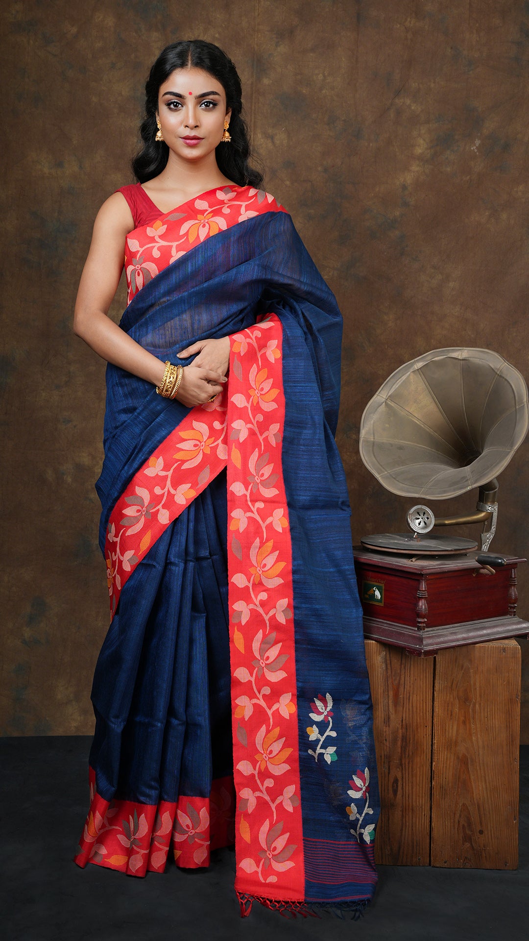 Deep blue raw silk saree with red silk border featuring beige, mustard, red and green woven pallu. Includes 0.8m running blouse piece. 5.5 meter tant dhakai weave